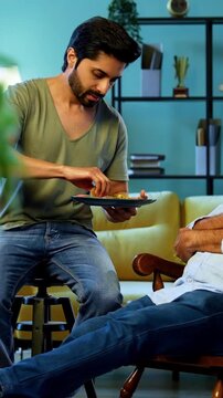 Indian young son feeding senior father chapati with sabzi from plate while he relaxes on chair, sharing heartfelt conversation and affection in modern home living room during homely mealtime