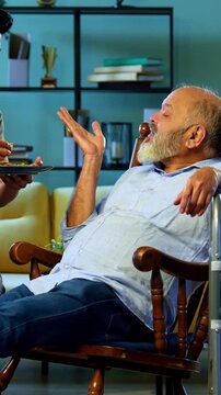 Indian young son feeding senior father chapati with sabzi from plate while he relaxes on chair, sharing heartfelt conversation and affection in modern home living room during homely mealtime