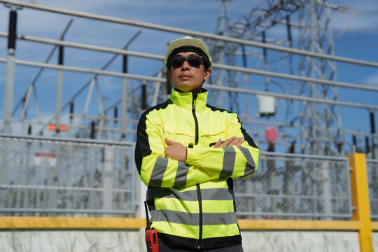 Asian electrical engineer standing with arms crossed, wearing hard hat and safety jacket, overseeing power grid infrastructure at a high voltage substation