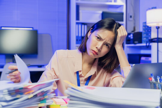 Stressed young Asian office worker feeling overwhelmed with paperwork and workload while working on laptop in office