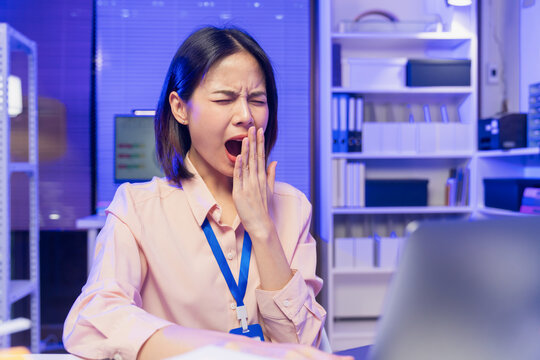 Tired young Asian office worker yawning while working on laptop in modern office.