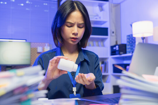 Young Asian businesswoman taking medicine from pill bottle while working in office, healthcare and wellness concept.