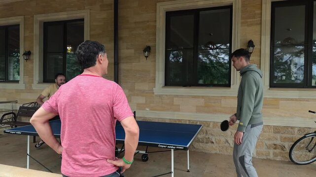 Family Men Playing Table Tennis on Covered Stone Terrace, Father Watching Son and Friend, Blue Outdoor Ping Pong Table, Red Paddles, Authentic Wide Shot in Soft Natural Daylight by Windows and Bicycle