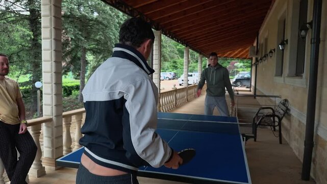 Father and son playing ping pong on covered veranda as family friend watches nearby, older man serving across blue table beside stone house wall, wooden roof and relaxed authentic leisure moment