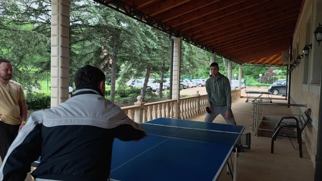 Father and son playing ping pong on covered veranda as son returns ball and older man serves, observer watching by railing during family leisure beside blue table, wooden roof and green trees outside