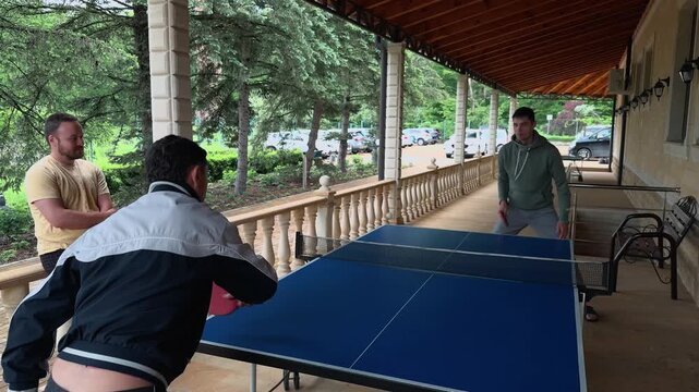 Father and son playing ping pong on covered veranda while friend watches by railing, older man serving toward young player near blue table, stone wall, wooden roof and trees in authentic family moment
