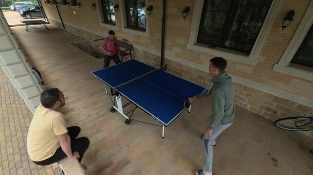 Overhead Men Playing Table Tennis on Covered Stone Terrace, Family Ping Pong Rally with Spectator on Railing, Blue Outdoor Table, Static Wide Angle View in Rainy Daylight by Windows and Bicycle