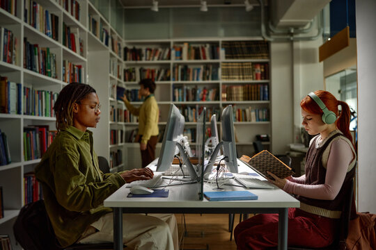 Biracial students studying in library, using desktop computers and reading book with headphones, supporting academic research and exam preparation
