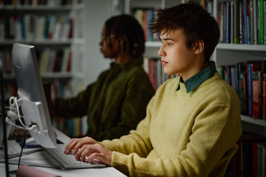 Male student using desktop computer in library, studying and researching coursework beside classmate
