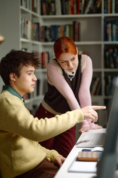Vertical shot of Caucasian students collaborating at computer, pointing at screen and discussing coursework in library

