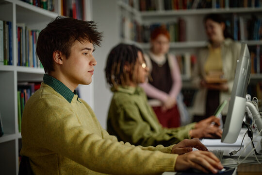 Male student using desktop computer in library, studying with classmates
