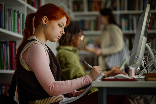 Woman studying at desktop computer, taking notes in library with classmates nearby. Education concept for academic success, exam prep, digital learning campaigns
