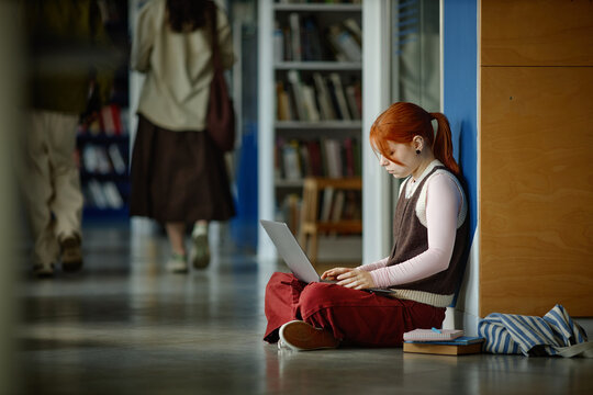 Student studying on laptop while sitting on library hallway floor near books, representing focused learning and digital education

