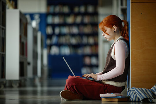 Female student studying on laptop while sitting on library floor, focusing on online learning and academic research
