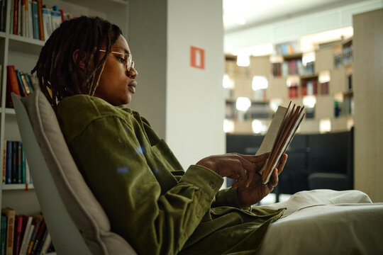 Young Black student reading book while sitting in library, studying independently and preparing for exams
