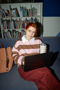 Portrait of Caucasian female student studying with laptop and takeaway coffee in library, using flexible learning concept. Useful for education marketing, campus life, e learning
