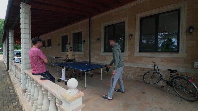 Wide Angle Family Table Tennis on Rainy Covered Stone Terrace, Father Watching Son and Friend Play Ping Pong, Blue Outdoor Table, Bicycle, Balusters, Authentic Leisure Shot in Soft Natural Daylight