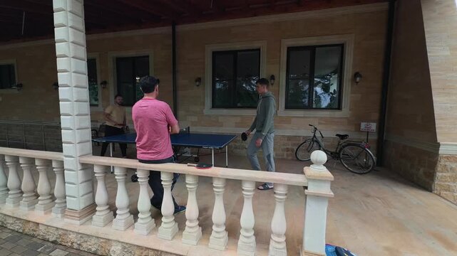 Wide Angle Men Playing Table Tennis on Rainy Covered Stone Terrace, Father Watching Son and Friend, Blue Ping Pong Table, Bicycle, Balusters, Authentic Family Lifestyle Shot in Soft Natural Daylight