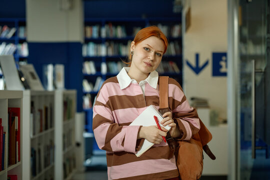 Portrait of Caucasian student holding notebook and pen, standing in library, preparing for study session and academic planning
