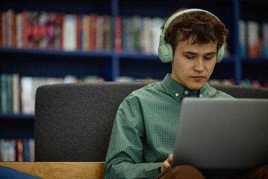 Young adult Caucasian male student wearing headphones, studying on laptop in library, focusing on online learning and academic research
