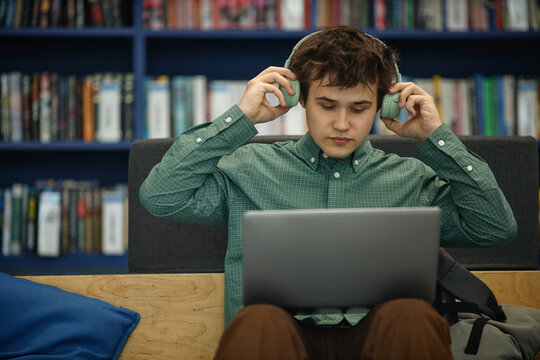 Male student adjusting headphones while studying on laptop in library, focusing on online learning and exam preparation
