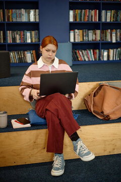 Vertical shot of Caucasian woman studying on laptop in library, sitting on steps with notebook and backpack. Useful for education marketing, e learning, student life content

