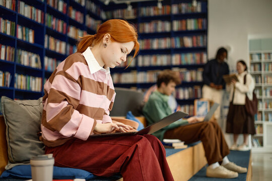 Young adult woman studying on laptop in library, focusing on coursework and digital learning among classmates. Education marketing, academic success, student life promotion
