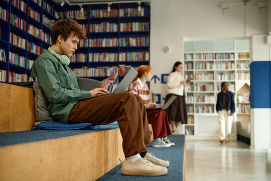 Male student studying on laptop in library, wearing headphones and preparing coursework while classmates learning nearby. Useful for education marketing, e learning, student life
