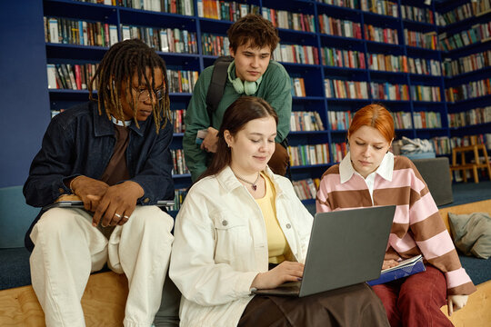 Young adult students collaborating on laptop research in library, sharing study materials and reviewing project together. Education marketing, academic teamwork, campus learning concept
