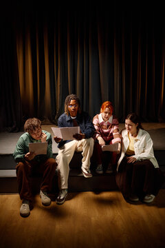 Vertical shot of diverse students reviewing scripts while sitting on stage steps, preparing school performance and teamwork project. Useful for education, youth theater, collaboration themes
