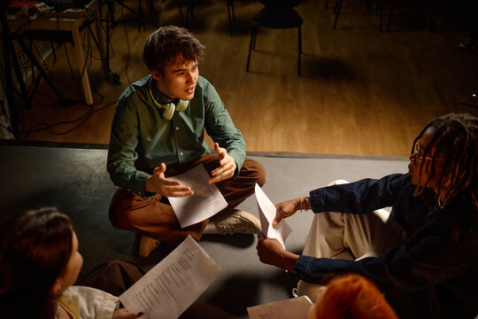 High angle shot of diverse students sitting on floor, discussing scripts during class project rehearsal
