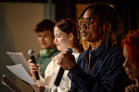 African American student speaking into microphone while classmates reading notes during school presentation, representing public speaking skills and education marketing use, close up
