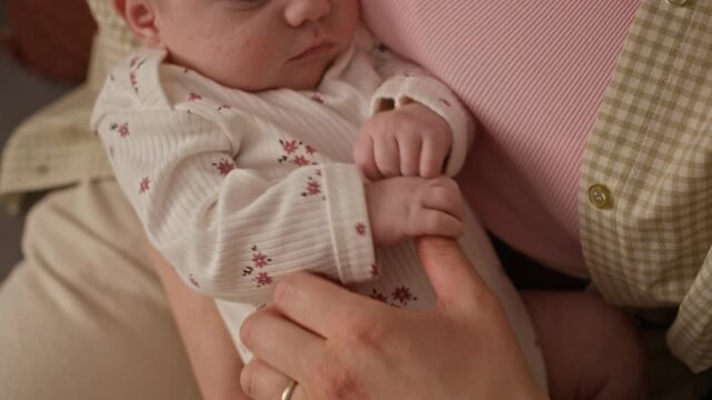 High angle shot of Caucasian newborn baby girl in soft white onesie with floral pattern grabbing unrecognizable mothers finger while lying in her caring arms at home