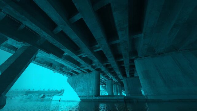 Concrete bridge showing structural beams and columns from an underground perspective, providing a symmetrical view of modern urban infrastructure over calm teal water