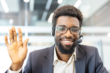 Obraz premium Professional african american call center agent wearing a headset and glasses, smiling while waving a hand, providing remote assistance and support from an office environment