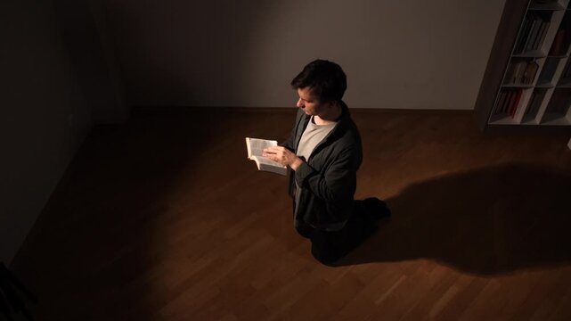 Young man kneeling on a wooden floor, deeply engaged in prayer and reading a holy book under a single beam of light, symbolizing faith, devotion, and spiritual reflection