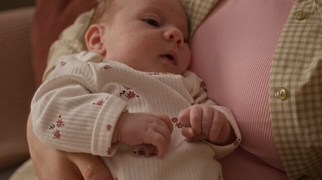 Slowmo shot of Caucasian infant baby girl in soft white onesie with floral pattern lying in caring arms of unrecognizable mother at home