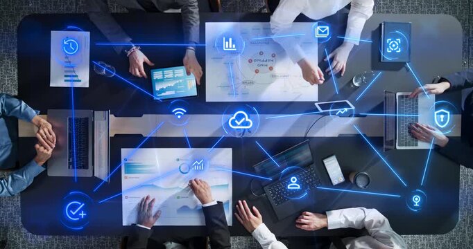 Top Down View of Business Team Working Around Office Table With Laptops, Tablets and Charts as Linked Visualization Layer of Icons Glowing Blue Cloud Connectivity, Cybersecurity, Data Analytics 