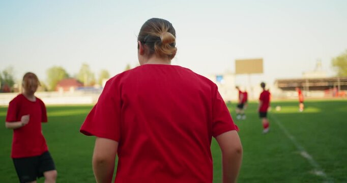 Two amateur soccer players in red jerseys practicing passes on a sunny outdoor grass field, with one athlete viewed from behind during training