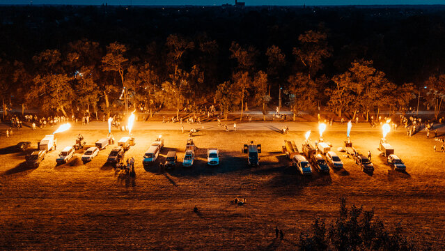 Aerial view of hot air balloon burners shooting flames into the night sky from a row of vehicles with crowds of people gathered in a park Senta, Vojvodina, Serbia.