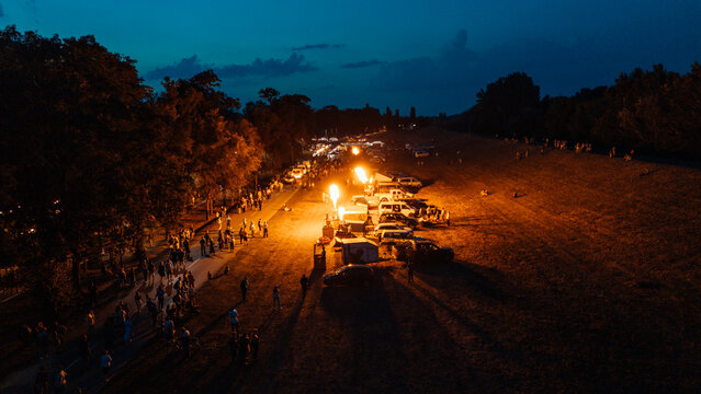 Aerial view of hot air balloon burners lighting up a night festival with crowds of people and parked vehicles in Senta, Vojvodina, Serbia.
