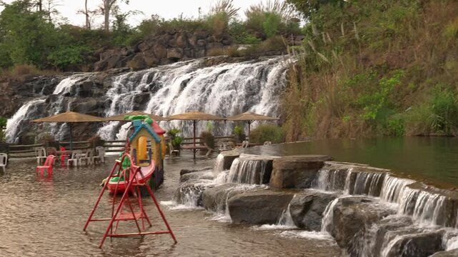 Static view of Tad Khoo waterfall in Laos, with cascades flowing behind a poolside recreation area with chairs, umbrellas and a small playground.