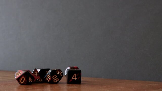 A pile of different dice like a D6, percentile dice, D4, D10, D12 and D8 sit on a wooden table. A player rolls a D20 into frame and after a few seconds picks it back up.