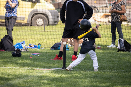 Young child swings bat at baseball field. Green grass covers entire playing surface. Adults stand behind netting, watching game. Equipment scattered nearby, including helmets.