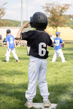 Young boy in black baseball jersey and helmet with white pants stands on base. Other players in blue jerseys stand nearby.
