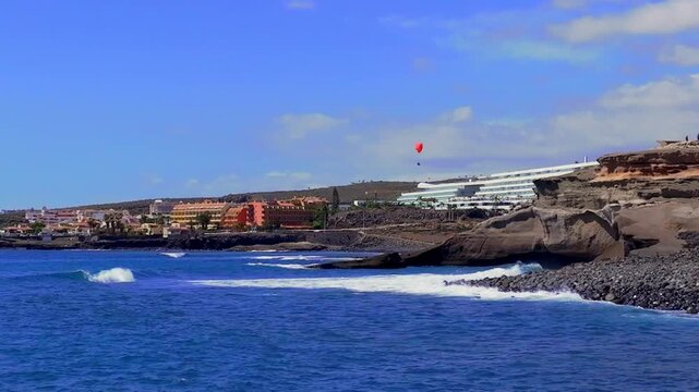 Paraglider glides over rocky coastline with waves near resorts on a sunny day