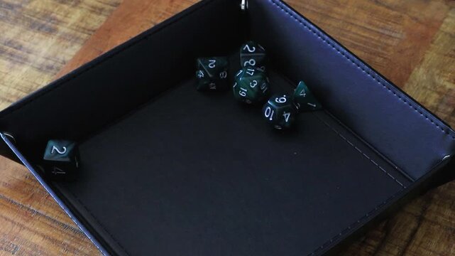 A player rolls a D20 into a black dice tray that sits on a wooden table. Inside of the dice tray there are already a few dice like a D12, D6, D10 and a D4