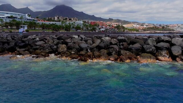 Waves hitting stone breakwater with resort hotels and palm trees in coastal Tenerife