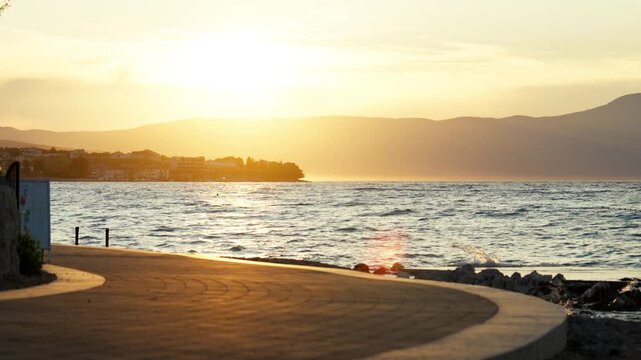 Warm sunset light over Adriatic Sea coastline on Krk island in Croatia with calm waves, seaside promenade and distant coastal town during summer evening.