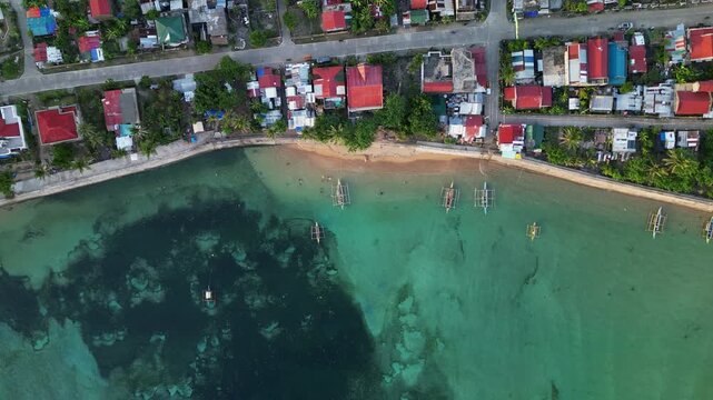 Aerial bird's eye pullback of traditional bangka fishing boats lined along quaint, coastal barangay town - Virac, Catanduanes, Philippines